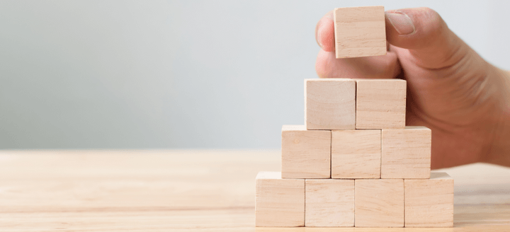 Hand placing a tiny wooden block on the top of a pyramid made of nine similar blocks standing on a wooden surface 