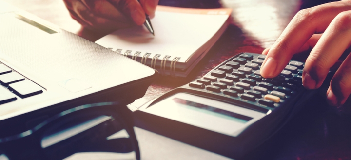 Pair of hands on a desk, with the left hand operating a desktop calculator and the right hand writing on a notepad with a pen