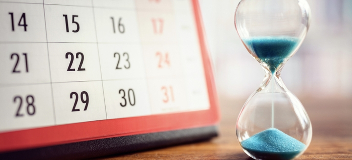 Sand timer with blue sand, next to and slightly in front of a desk calendar on a desktop