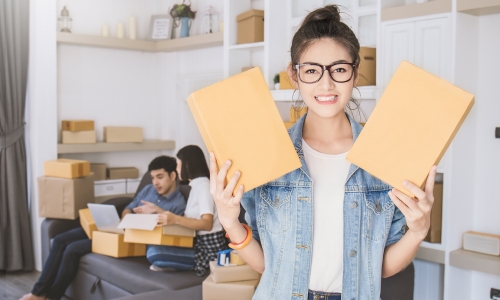 Young woman holding two boxes and two people working while sitting on a couch in the background